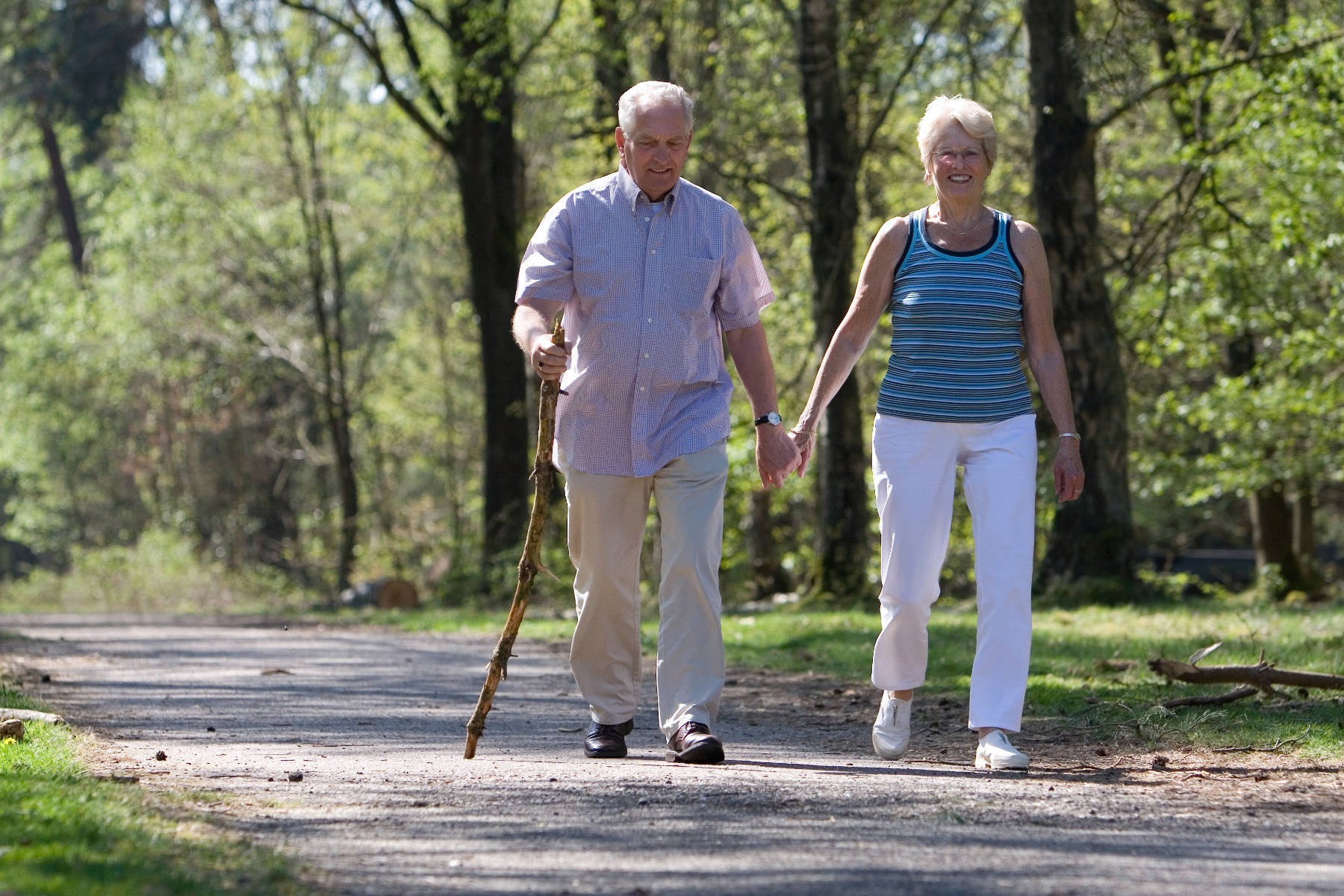 A couple in their 50s walking in the forest, hand-in-hand, the man has a walking stick.